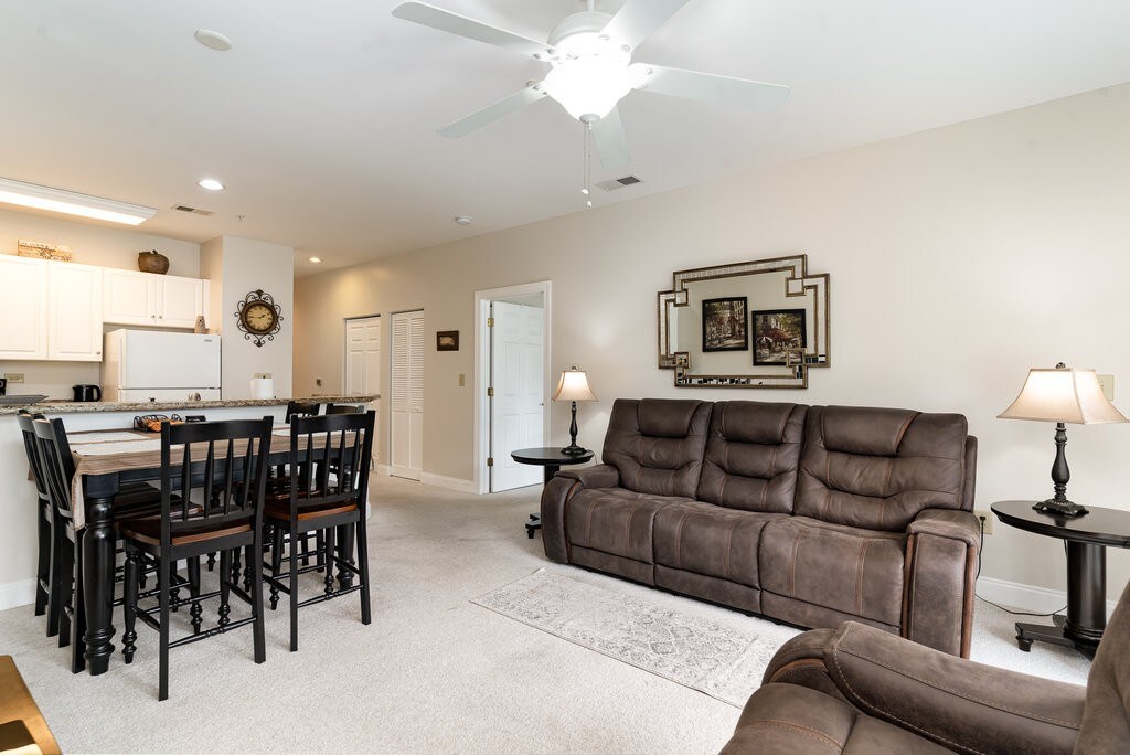 A spacious living area features a comfortable brown reclining sofa complemented by two side tables with lamps. A dining table with high-backed chairs is visible in the background, while a kitchen area can be seen through an open doorway. Soft lighting enhances the relaxed ambiance.