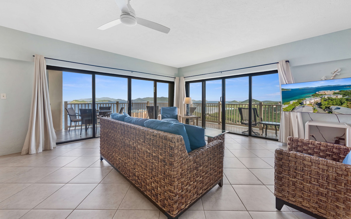 A spacious living area features large glass doors that provide sweeping views of the hills and ocean. Comfortable wicker furniture, accented with blue cushions, is arranged thoughtfully. A ceiling fan circulates air while a television stands to one side, enhancing the relaxation space.