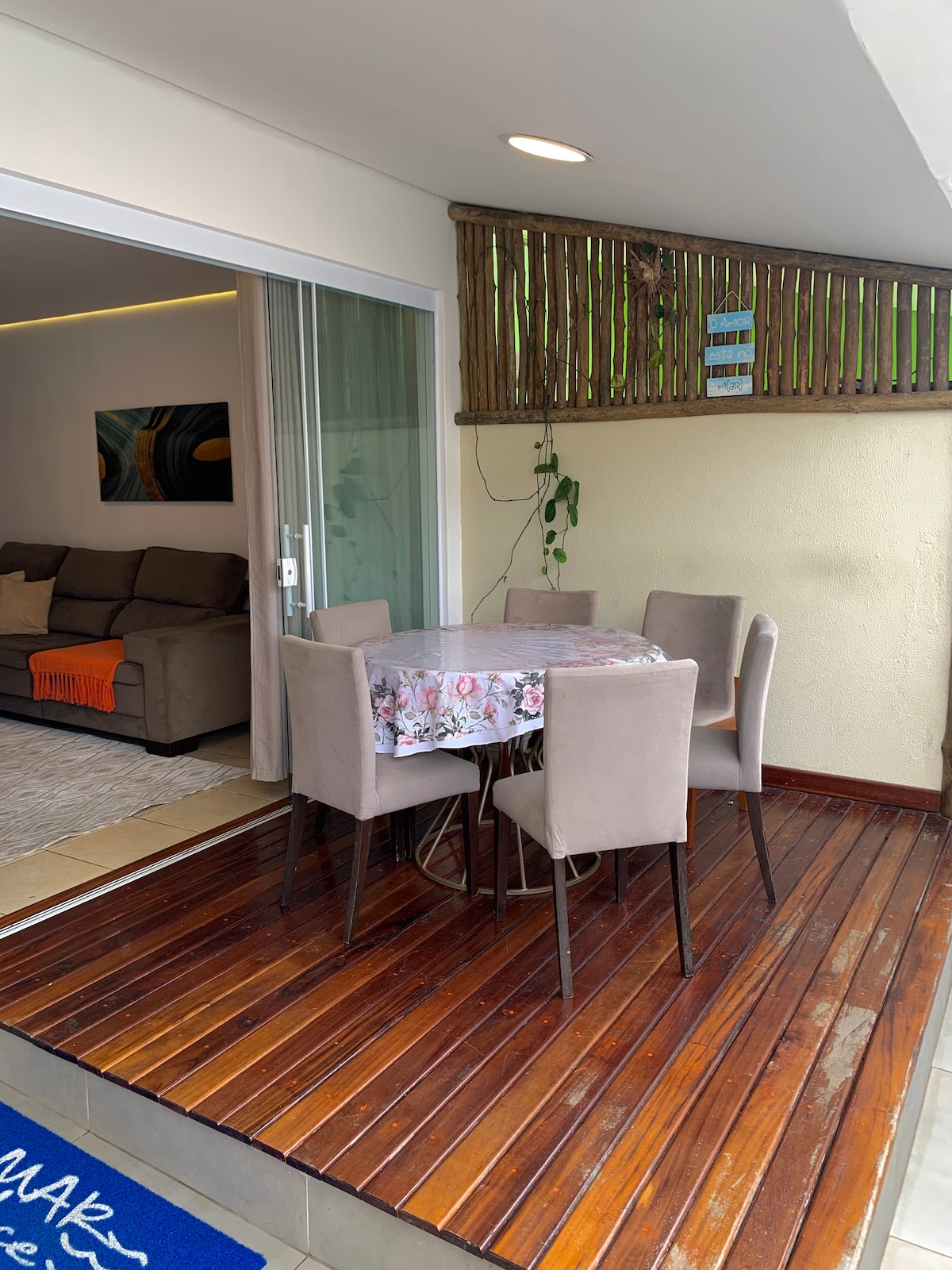 An outdoor dining area is set on a wooden deck, featuring a square table covered with a floral tablecloth. Surrounding the table are upholstered chairs. In the background, a cozy living area can be glimpsed through sliding glass doors, adding to the inviting space.