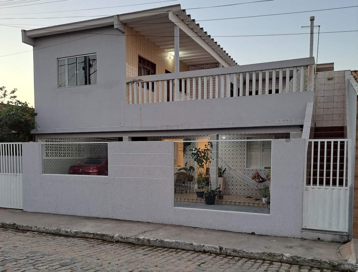 The exterior of a two-story house is displayed, featuring a white facade and a balcony on the upper level. A large window showcases the interior space, where plants are visible. A fenced area includes a driveway with a parked car.