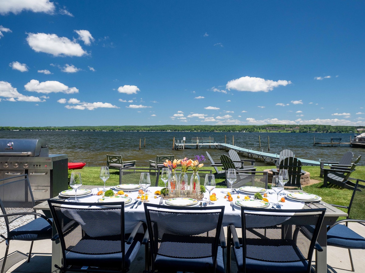 A dining setup is showcased on a patio facing the lake, featuring a long table adorned with flowers and dishes. Grassy areas with adirondack chairs and a grill complement the picturesque lake view under a bright blue sky with fluffy clouds.
