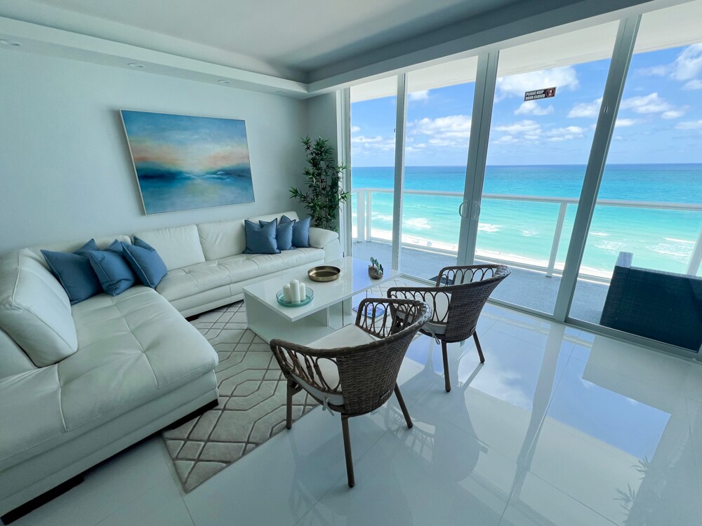 A bright living area features large glass doors that open to a stunning ocean view. A white sectional sofa is decorated with blue pillows. A glass coffee table sits in the center, accompanied by two woven chairs. The light reflects off the glossy tile floor, enhancing the airy atmosphere.