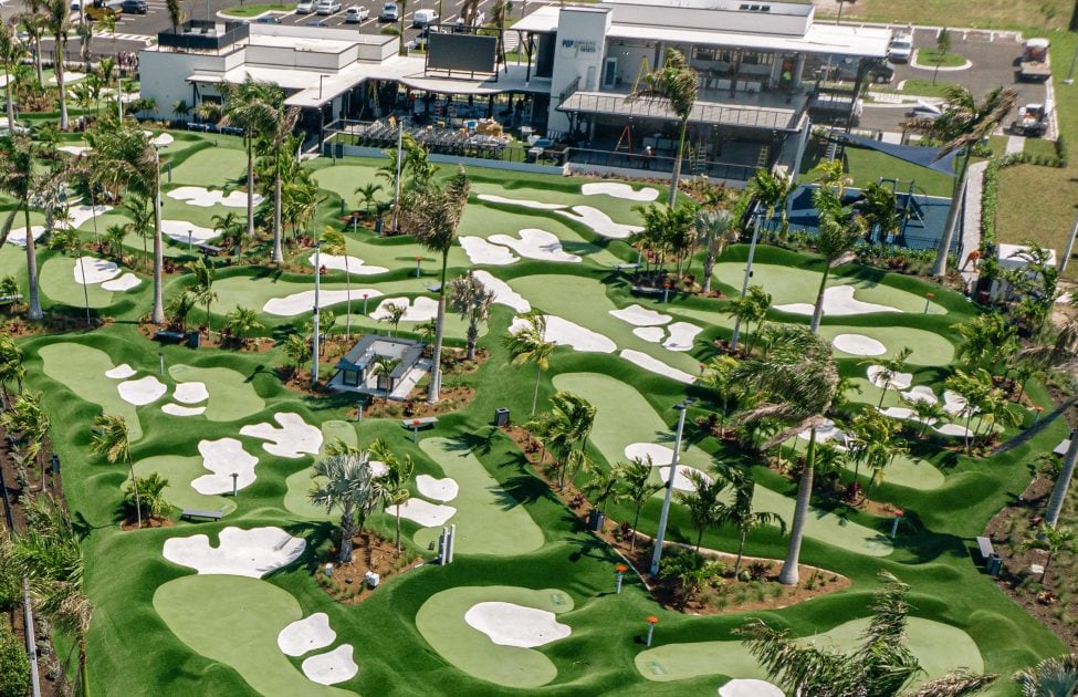 An aerial view of a mini golf course features multiple grassy greens intricately designed with white sand traps scattered throughout. The landscape is enhanced by surrounding palm trees and a modern building nearby, providing a vibrant recreational atmosphere.