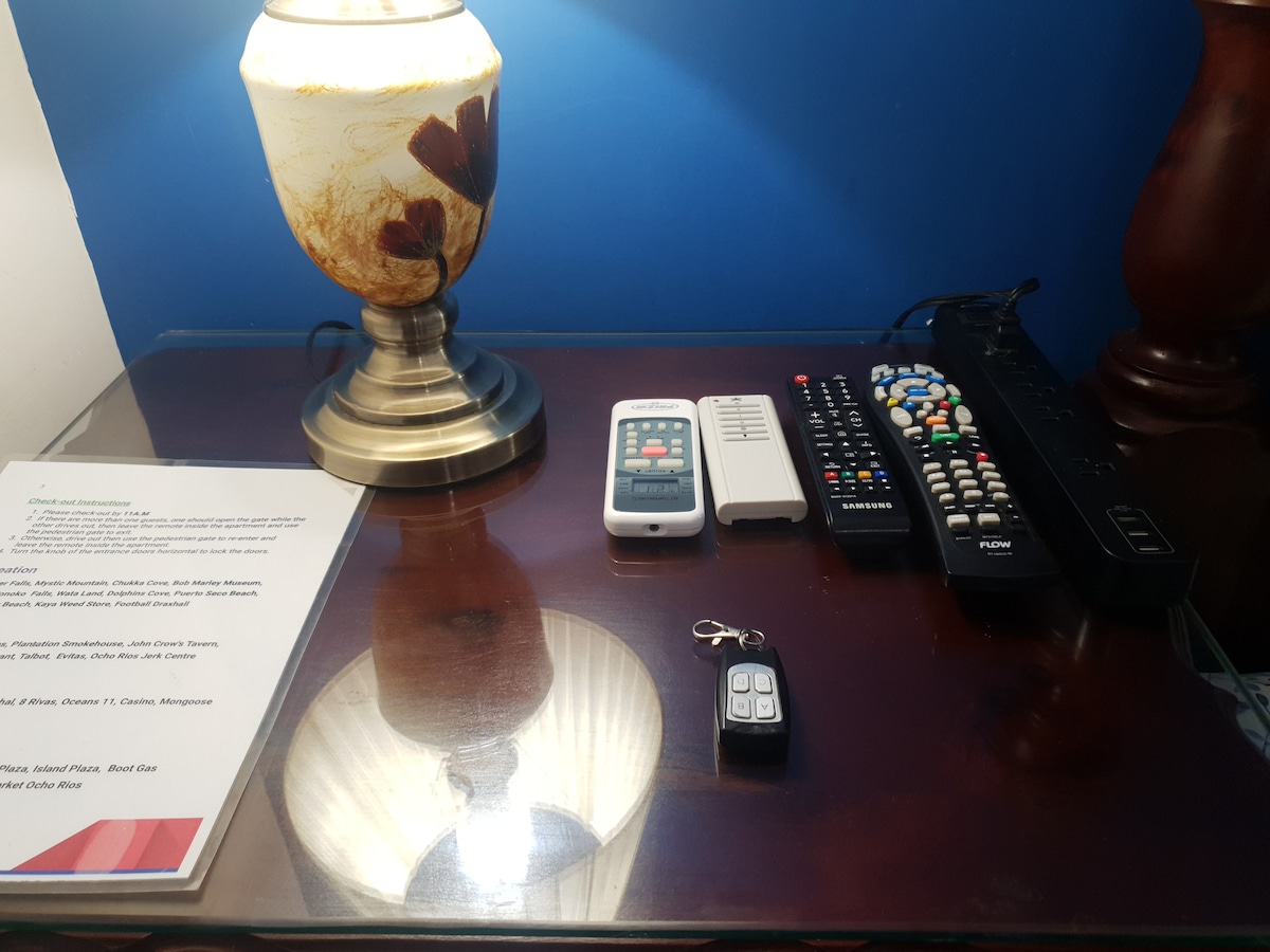 A bedside table is shown, featuring a warmly designed lamp with a decorative shade. Various remote controls are arranged alongside a small device, a guest information booklet, and a security remote. The glossy surface reflects the lamp and surrounding items.