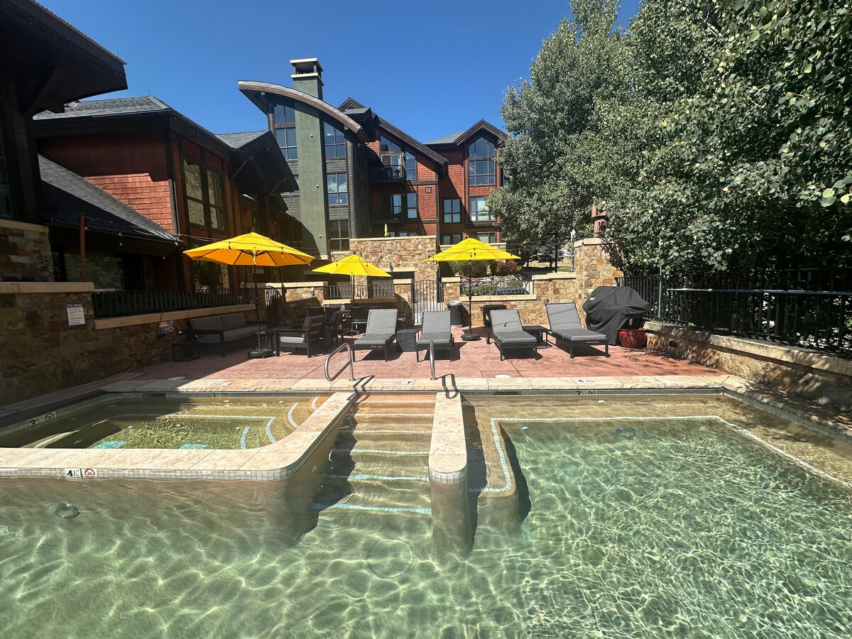 A serene outdoor space is highlighted by a heated pool, designed with stone steps leading into clear waters. Lounge chairs are arranged around the pool, accompanied by vibrant yellow umbrellas providing shade. A gas grill and lush greenery are visible in the background.