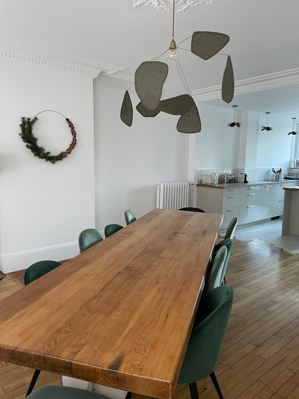 A long wooden dining table is surrounded by green upholstered chairs, creating a welcoming dining area. A unique pendant light hangs above the table, and a wreath is displayed on the wall. The open layout connects to a well-equipped kitchen in the background.