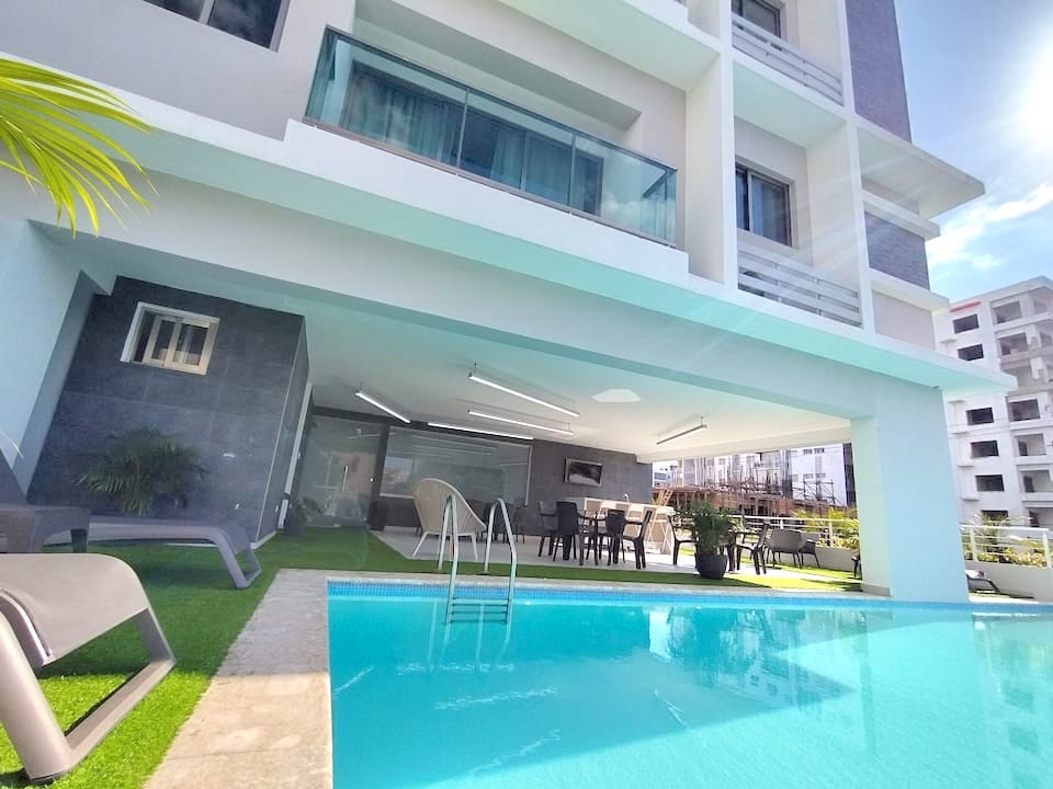An outdoor area features a clear swimming pool, bordered by a green lawn and sun loungers. A spacious covered patio is visible, furnished with a dining table and chairs, providing a comfortable space for gatherings, while large glass doors offer a view of the interior.