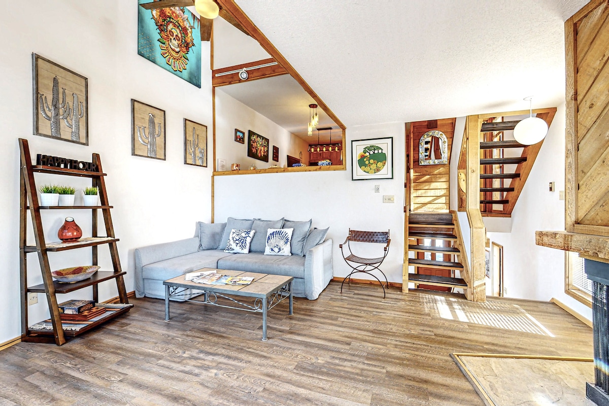 A bright living area features a light-colored couch adorned with decorative pillows. A coffee table sits in front, accompanied by a shelving unit displaying artwork and decorative items. The space is complemented by artwork on the walls and a staircase leading to the upper levels.