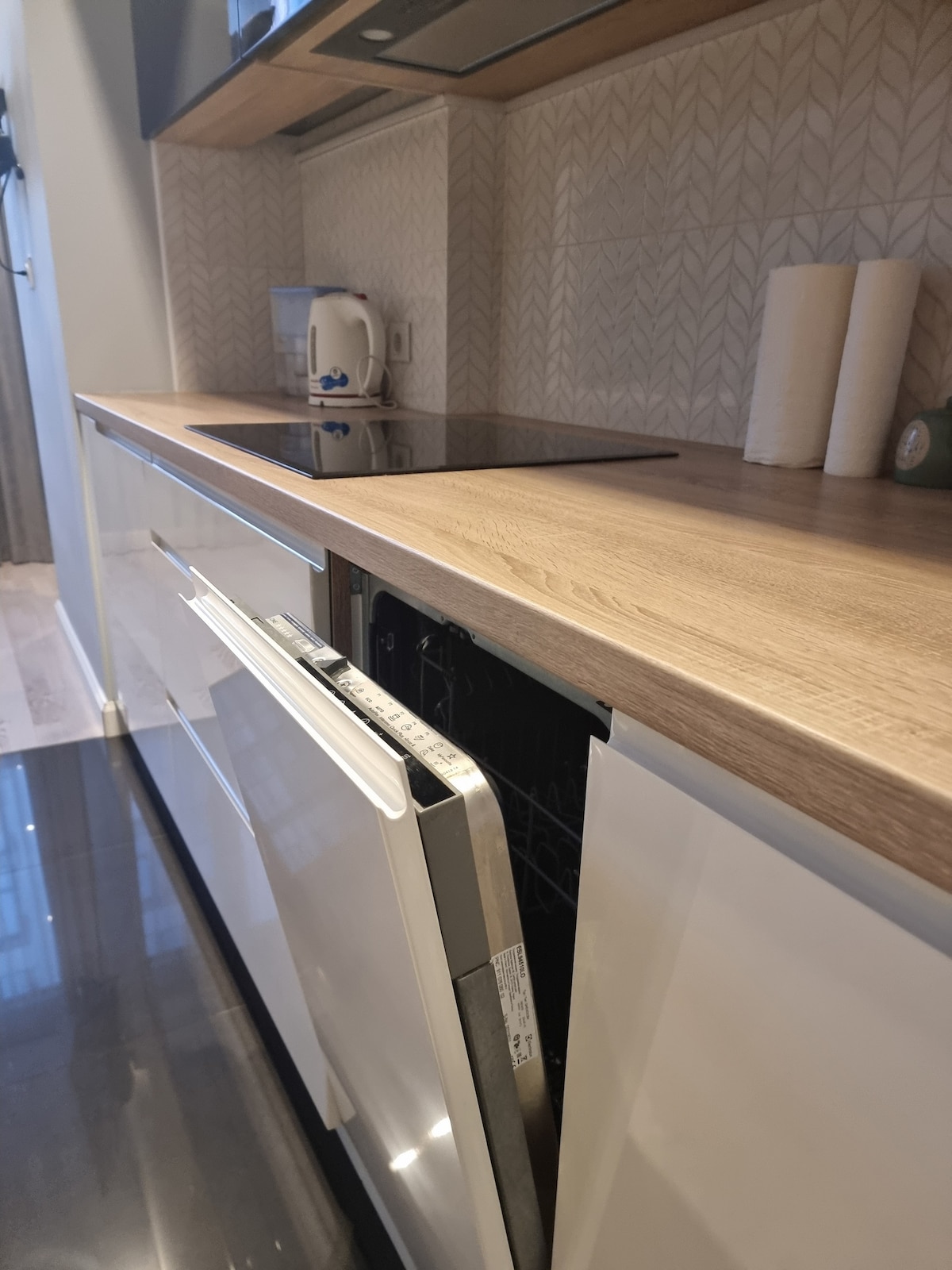 A modern kitchen area is featured, with sleek white cabinets and a wooden countertop. An open dishwasher is visible, revealing its silver interior. The backsplash is decorated with a subtle chevron pattern, while kitchen essentials such as a kettle and paper towels are neatly arranged on the counter.