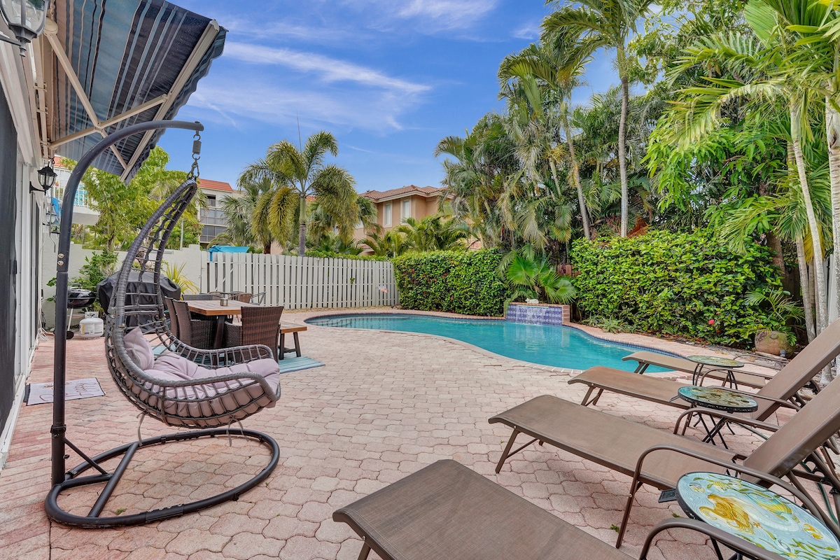 The outdoor area features a refreshing swimming pool surrounded by palm trees. Comfortable lounge chairs are arranged around the pool, and a hanging chair adds a cozy spot for relaxation. A dining table under a sunshade provides a space for outdoor meals.