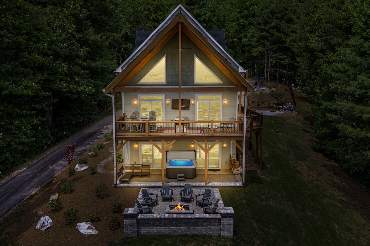The cabin's exterior showcases a three-story structure with large glass doors and windows, providing a view of the inviting living spaces inside. A spacious fire pit patio with comfortable seating is placed in front, surrounded by natural landscaping and trees.