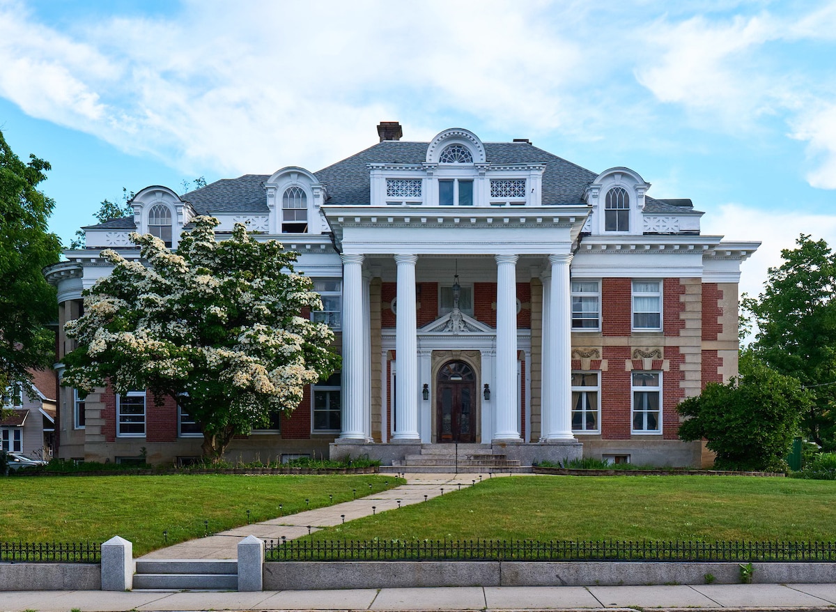 The historic Wheeler Mansion displays a grand façade featuring white columns and a large, arched entryway. Lush greenery is visible in front, with a flowering tree and a manicured lawn leading up to the entrance. The building showcases a mix of brick and decorative trim.