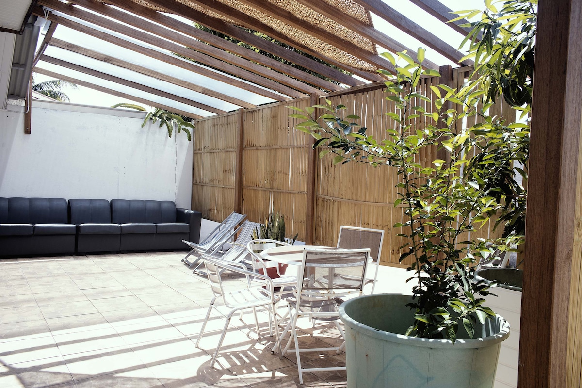 An outdoor terrace features a combination of a shaded seating area with a dark sofa and several sun loungers positioned on tiled flooring. A large potted plant adds a touch of greenery, while bamboo fencing provides privacy and a natural element to the space.