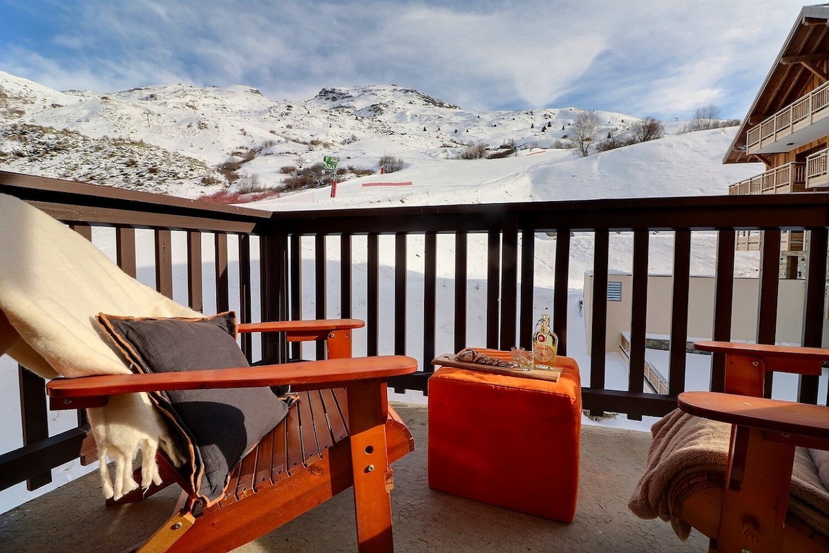 A private balcony is furnished with two wooden lounge chairs and a small orange table. A cozy blanket is draped over one chair. A snowy mountain landscape stretches in the background, providing a serene winter setting.