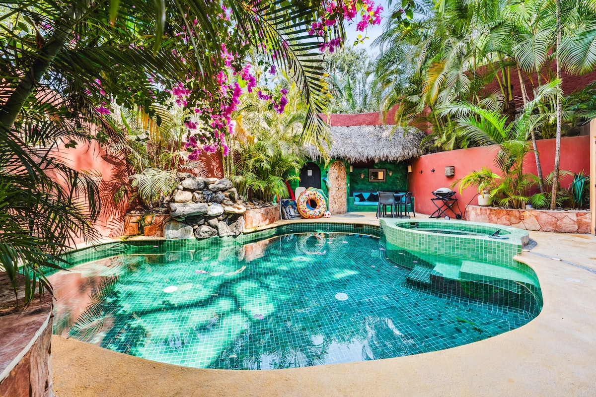 A private pool is surrounded by lush tropical foliage and vibrant flowers. The pool features a cascading stone fountain and an attached hot tub, all set against earthy toned walls. Comfortable seating areas are positioned nearby for relaxation.