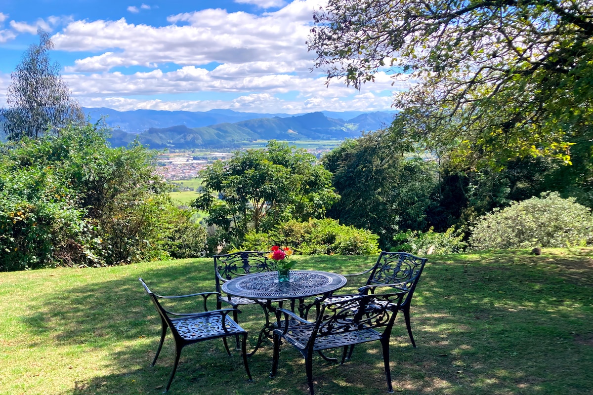 An outdoor seating area features a round table surrounded by four chairs, set against a backdrop of verdant trees and expansive mountain views. A small vase with flowers adorns the table, enhancing the serene atmosphere of the natural surroundings.