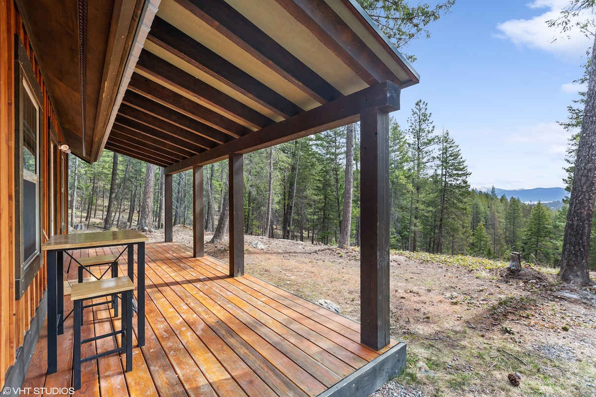 An inviting porch is showcased with a rustic wooden table and chair set. The expansive wooden deck offers a view of the surrounding trees and distant mountains under a clear blue sky.