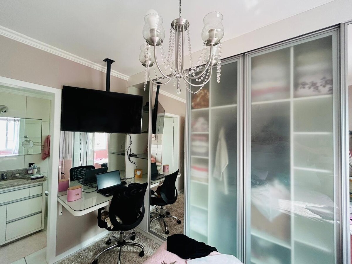 A well-lit workspace is showcased, featuring a desk with a computer and a stylish chair. A large wall-mounted TV is visible above the desk. The room is enhanced by a modern chandelier and mirrored elements, while the closet door displays frosted panels.