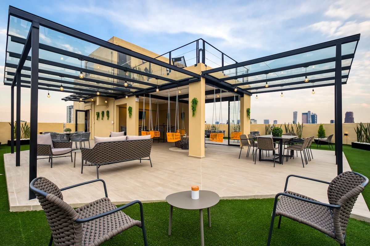 A spacious rooftop area features modern lounge seating on a grassy surface, with elegant swings and dining tables under a transparent canopy. String lights are draped overhead, creating a relaxed ambiance with views of the city skyline in the background.