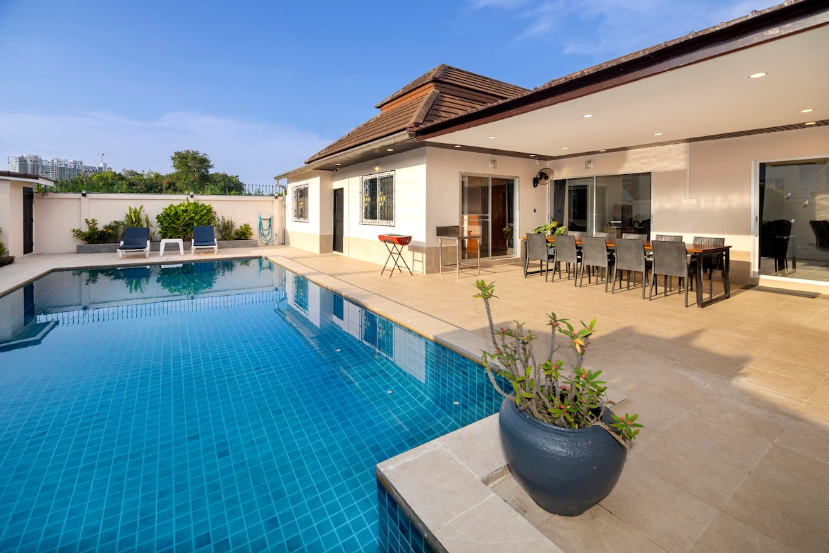 The image captures a large private swimming pool surrounded by a sun-drenched terrace. A dining area with a table and chairs is positioned next to the pool, with a barbecue grill visible. Lush greenery adds a touch of nature to the space, complemented by the villa's modern architecture.