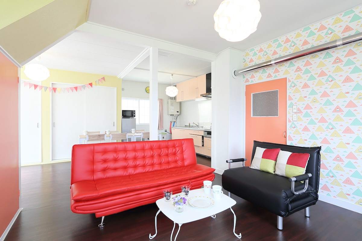 A modern living area features a vibrant red sofa and a sleek black armchair, complemented by a white coffee table. The space is brightly lit, showcasing colorful wall patterns and a light, airy atmosphere with an open layout that connects to the kitchen.