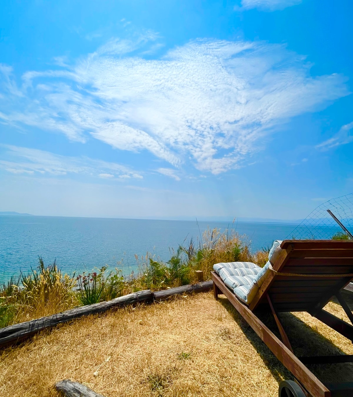 A wooden lounge chair sits on a grassy area overlooking the calm ocean. The sky is bright blue with wispy clouds, framing a serene view of the water. Sunlight reflects off the surface, creating a peaceful atmosphere.