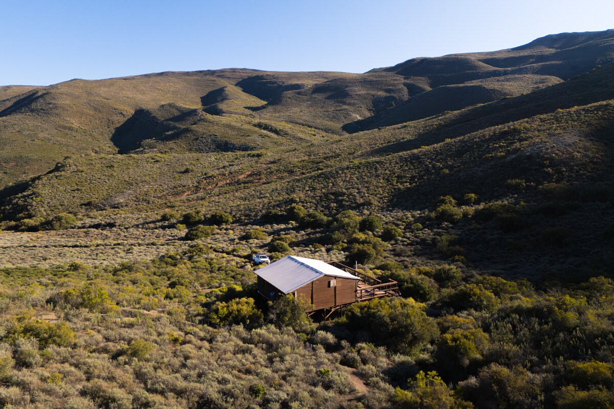 A wooden cabin is nestled amidst rolling hills and diverse vegetation. The structure is surrounded by native flora, set in a mountainous landscape that showcases the natural beauty of the Western Klein Karoo. The sunlight illuminates the scene, enhancing the tranquil atmosphere.