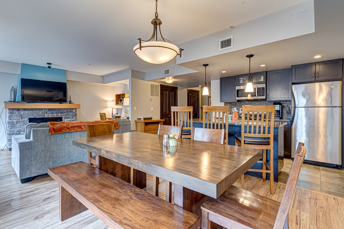 A spacious dining area features a large wooden table surrounded by matching chairs and a bench. An adjacent kitchen area is visible, with modern appliances and light fixtures contributing to a bright, open atmosphere. An electric fireplace can be seen in the living area.