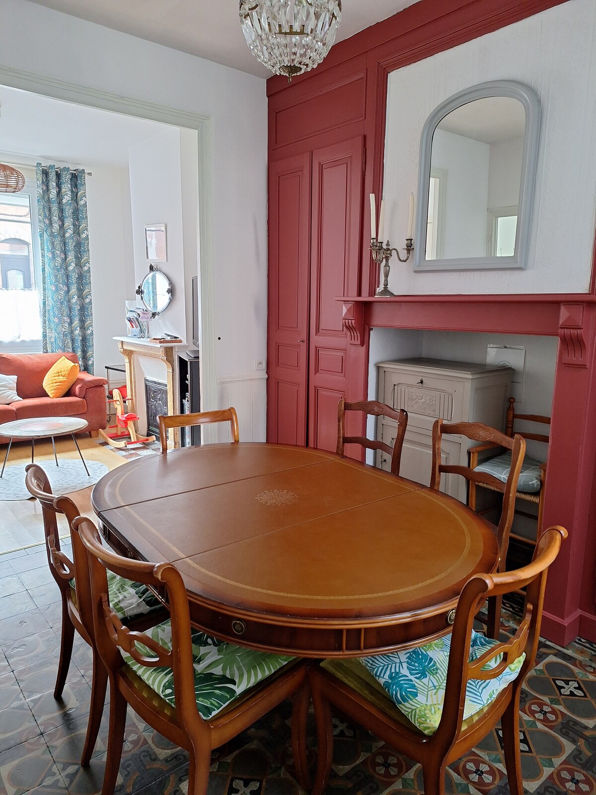 A round wooden dining table is surrounded by eight wooden chairs with colorful cushions. The wall behind features a decorative mirror and a storage cabinet. Soft light filters in from an adjacent room, highlighting the stylish decor.