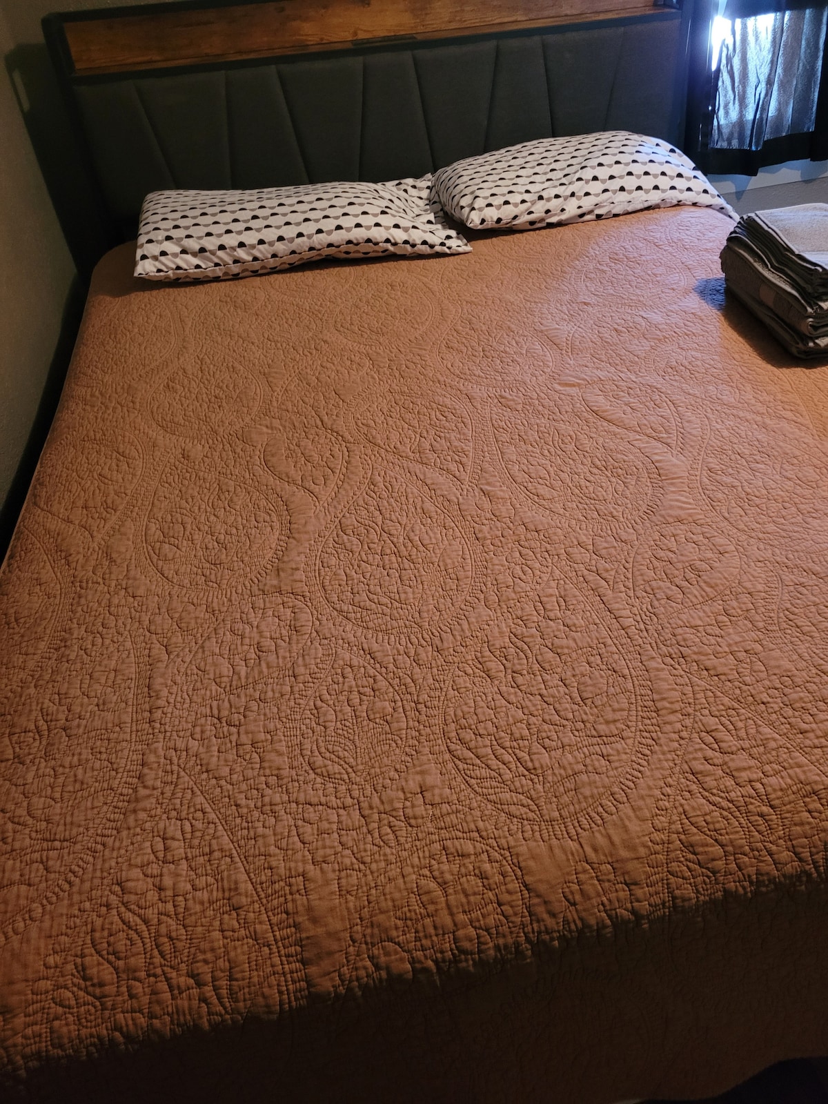 A neatly made Queen-sized bed is featured, with a textured orange quilt laid across it. Two patterned pillows are positioned at the head of the bed, with a folded blanket resting nearby. Soft light filters in from a partially covered window.