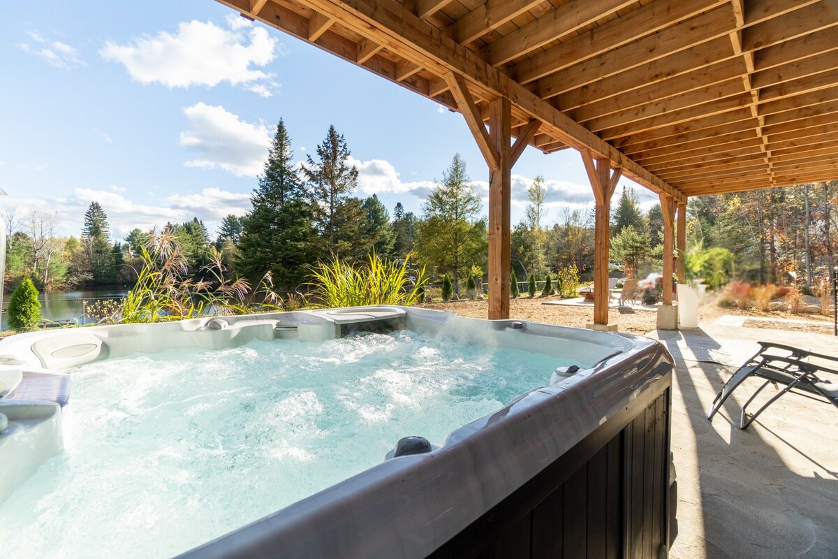 A year-round Sundance spa is positioned under a wooden structure, surrounded by greenery and tall trees. Sunlight filters through the open area, illuminating the bubbling water. Spacious seating is available nearby, creating a relaxing outdoor space.