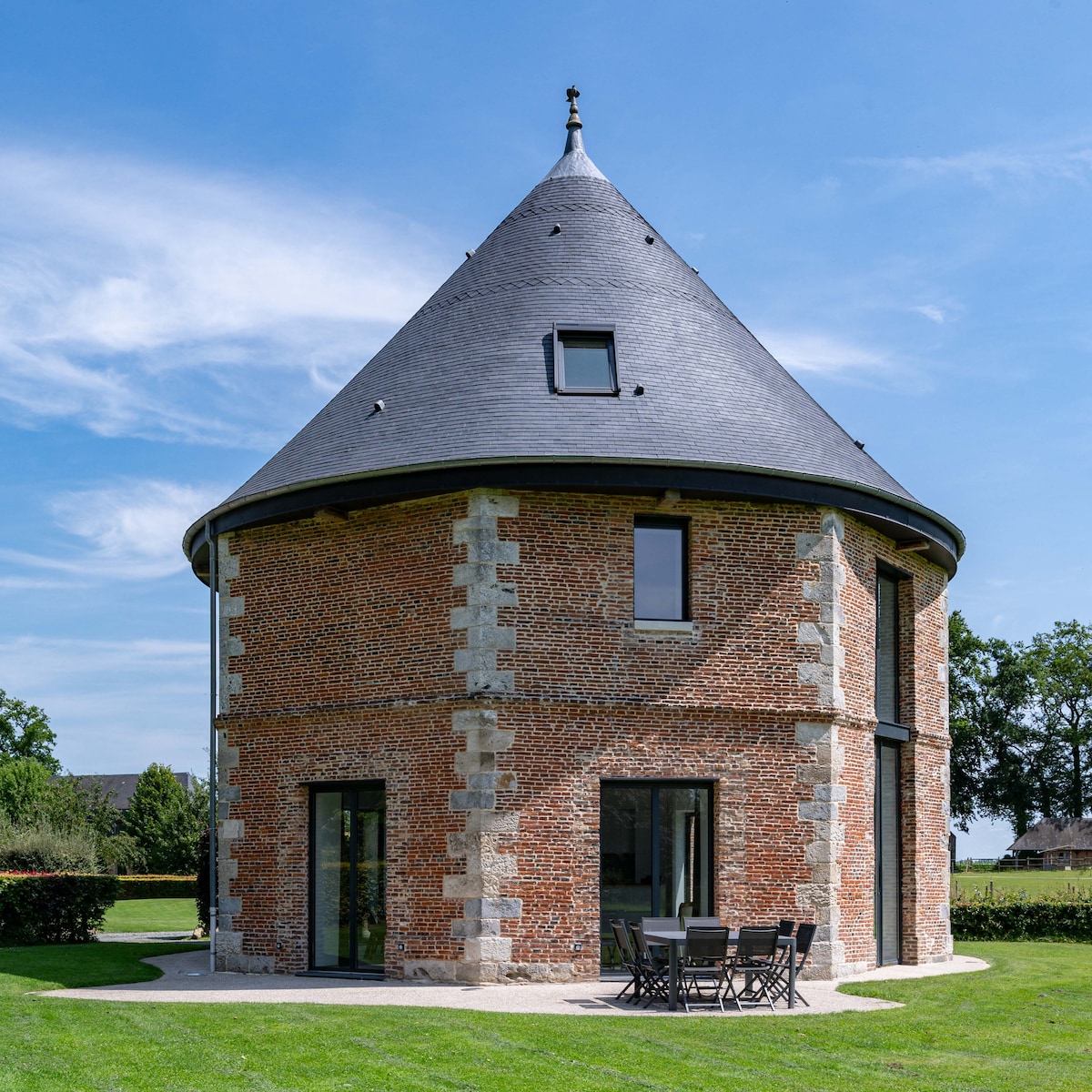 The exterior of the historic octagonal brick building features a prominent conical roof and large windows. A circular patio area with a black table and chairs is positioned on the grassy lawn, surrounded by lush greenery, offering a serene outdoor setting.