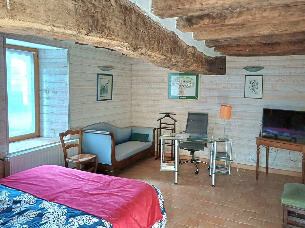 A spacious room features exposed wooden beams and a large window allowing natural light. A cozy bed with a floral-patterned cover faces a glass desk and chair setup. Seating options include a blue sofa and a wooden side table, contributing to a relaxed atmosphere.