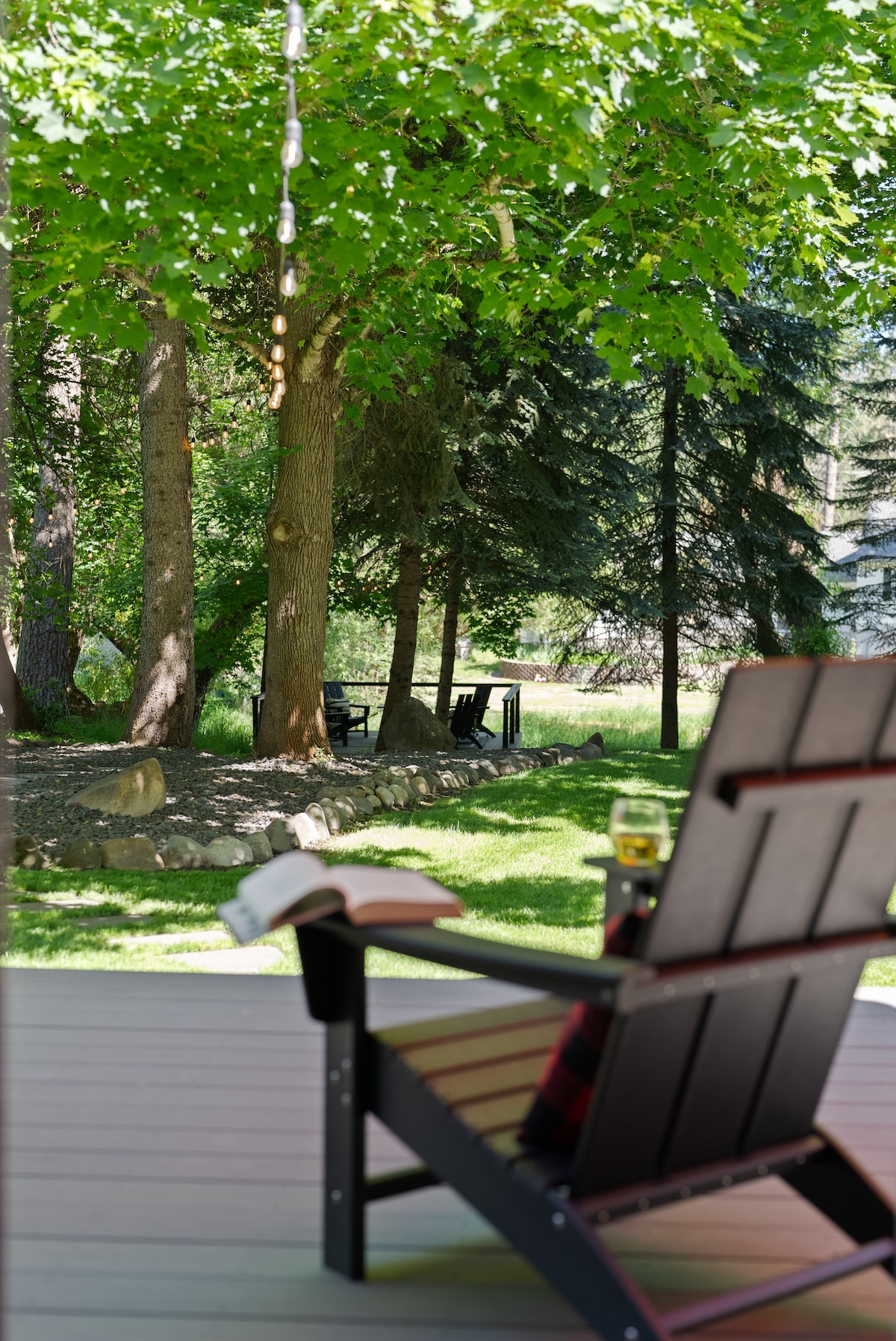 A single Adirondack chair is positioned on a shaded porch, offering a view of a vibrant green lawn adorned with trees. A book rests on the arm of the chair, while a glass is placed nearby, suggesting a serene outdoor setting.