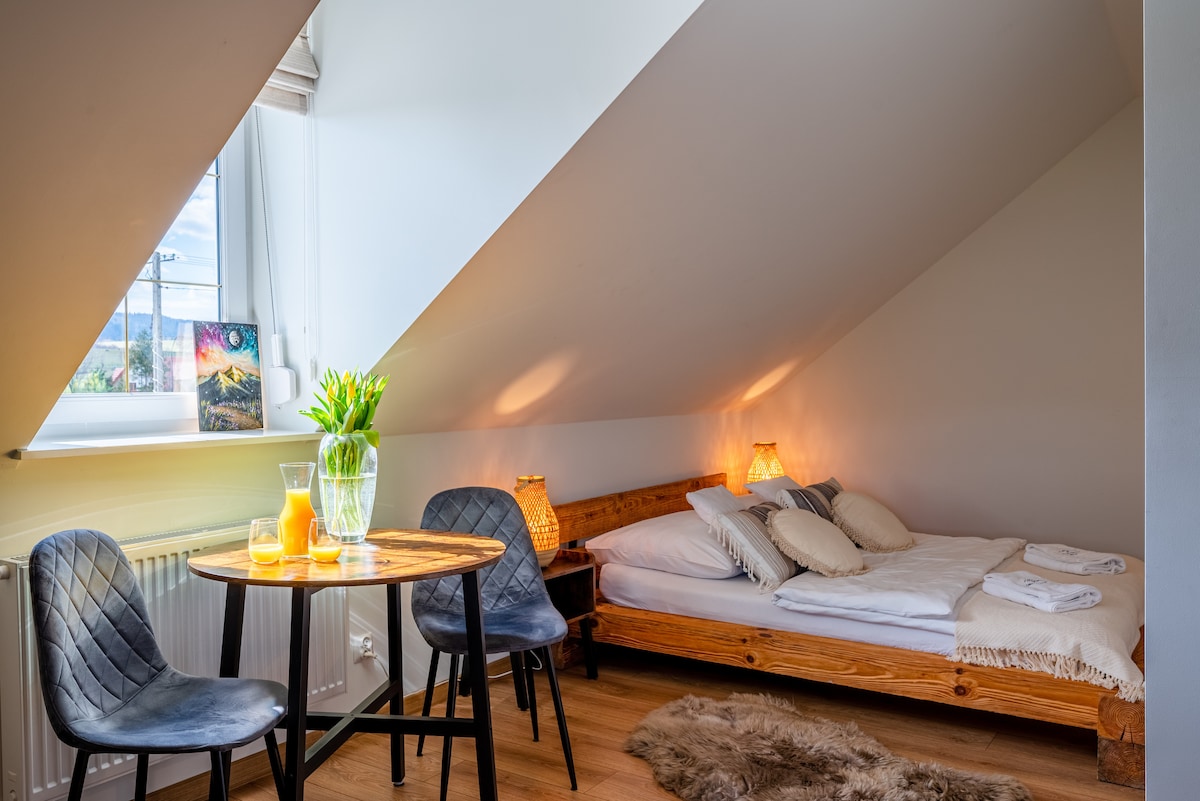 A cozy bedroom features slanted ceilings and natural light from a window. A wooden bed is dressed with soft linens and cushions. A small dining table with two chairs rests beside a decorative vase containing fresh flowers, providing a homely touch.