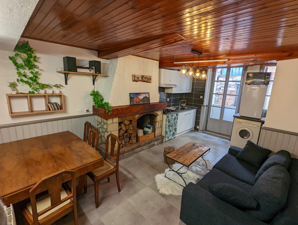 A spacious living area features a wooden dining table surrounded by chairs. A cozy seating area includes a dark sofa and a coffee table. A well-equipped kitchen is visible, complemented by a central fireplace with stacked wood. Natural light enters through the windows.