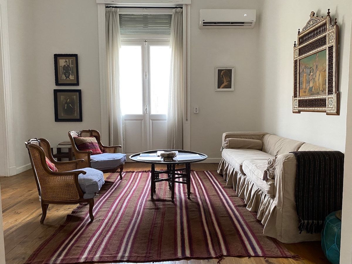 A spacious living room features a mix of classic and modern furnishings. Two armchairs and a sofa are arranged around a circular coffee table, with a colorful striped area rug underfoot. Light streams in through large windows, framed by sheer curtains.