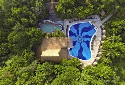 An aerial view showcases a large, curvy swimming pool with a vibrant blue design surrounded by lush green foliage. A separate, circular hot tub lies adjacent to the pool. Thatched-roof cabanas provide shaded seating areas along the pool's edge.