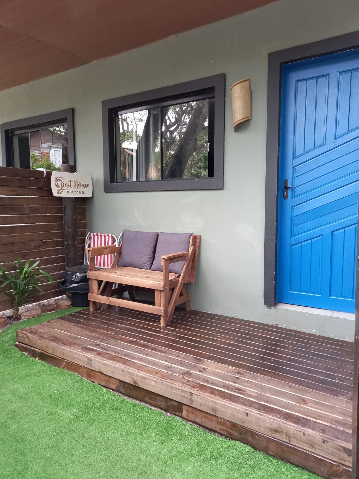 An entryway features a bright blue door and a wooden deck with a small seating area. A wooden lounge chair is positioned on a grassy surface, accompanied by a striped cushion. The surrounding walls are painted in a muted green, framed by dark window trims.