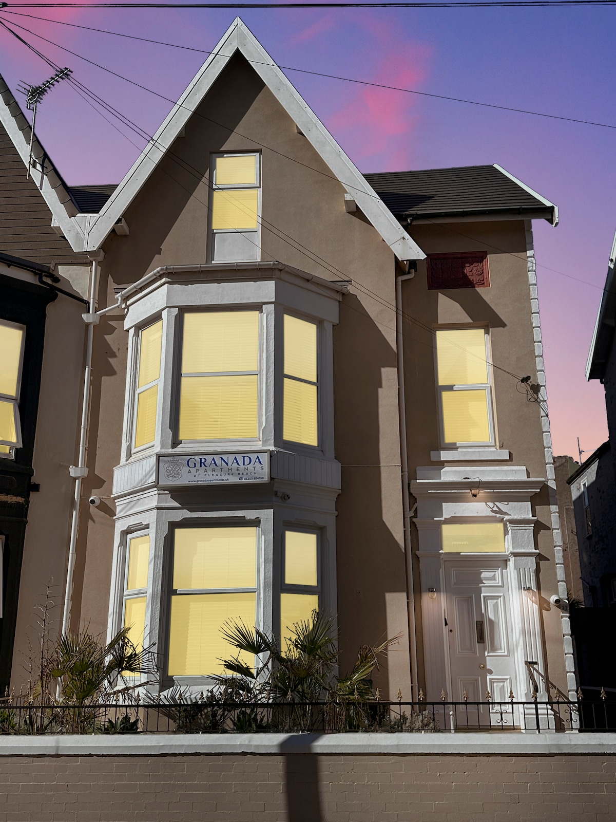 A stately building is viewed from the exterior, showcasing a Victorian architectural style. Large bay windows with yellow-tinted panes allow for plenty of natural light. Decorative plants are positioned in the front garden, contributing to the overall inviting appearance of the property.