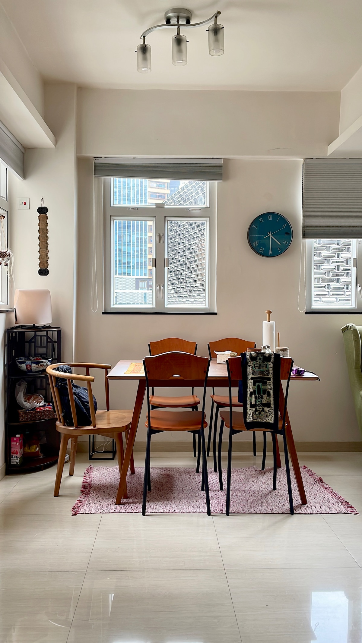 A dining area is presented with a wooden table surrounded by four chairs. Natural light enters through two windows, illuminating the space. A rug lies beneath the table, and decorative items are positioned on a nearby shelving unit.