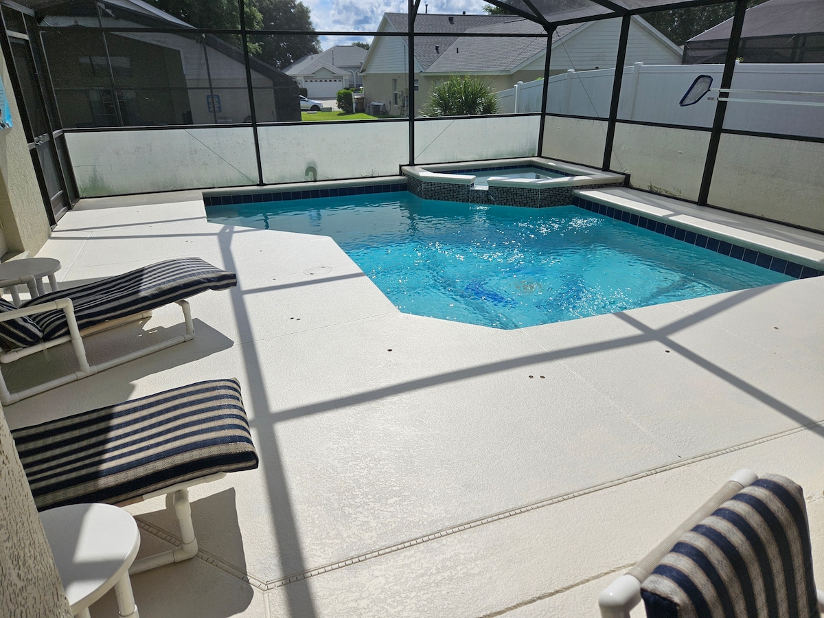 A private pool area features a crystal-clear swimming pool and adjacent spa, surrounded by a screened enclosure. Two lounge chairs with striped fabric are positioned for relaxation, with a spacious deck allowing for comfortable gatherings. Sunlight reflects off the water, creating a serene atmosphere.