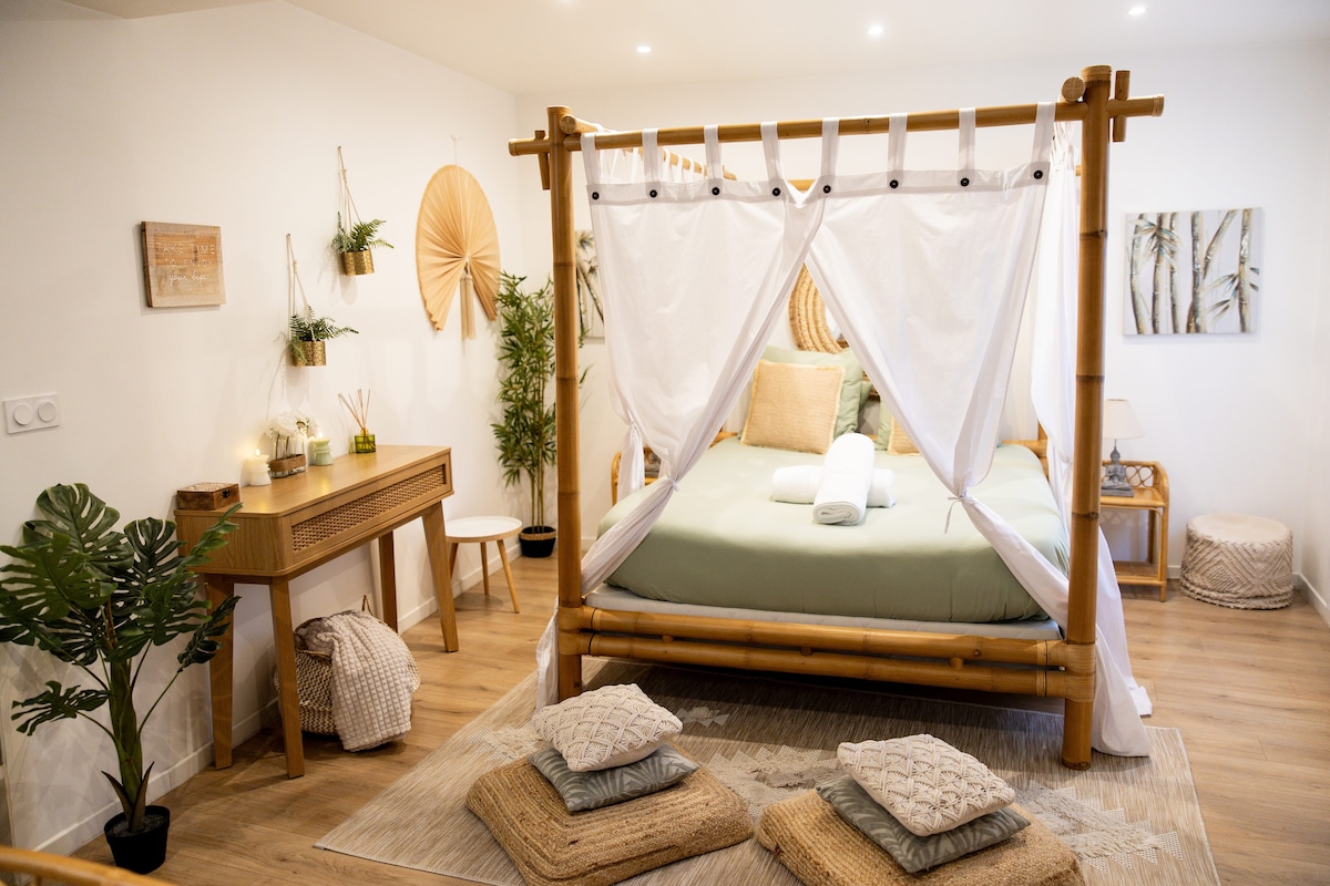 A spacious suite features a bamboo four-poster bed, draped in white fabric. An inviting green bedding is complemented by soft, textured cushions on the bed and surrounding floor. A small workspace and decorative elements, including plants and wall art, enhance the calming atmosphere.