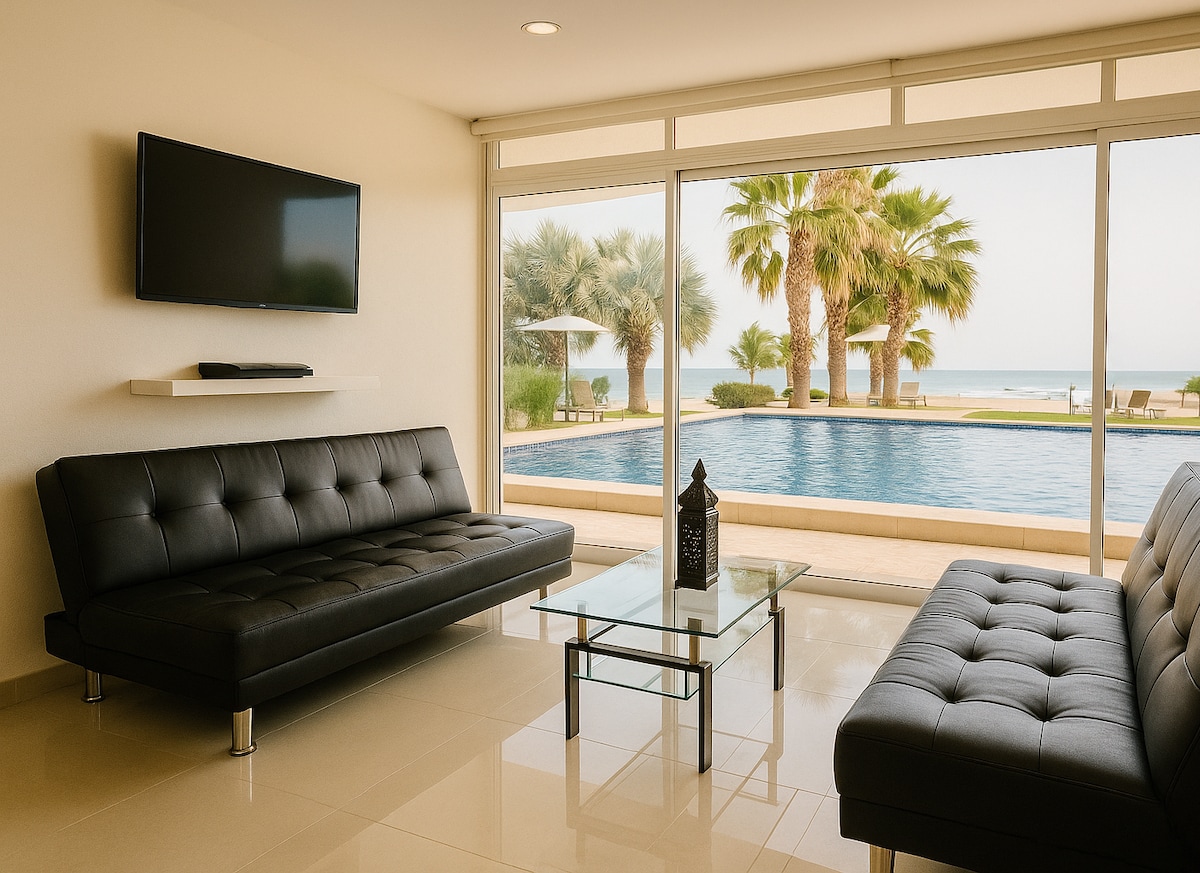 A modern living area is presented with two sleek black sofas facing a glass coffee table. Large windows frame a view of the pool and palm trees outside while allowing natural light to illuminate the room.