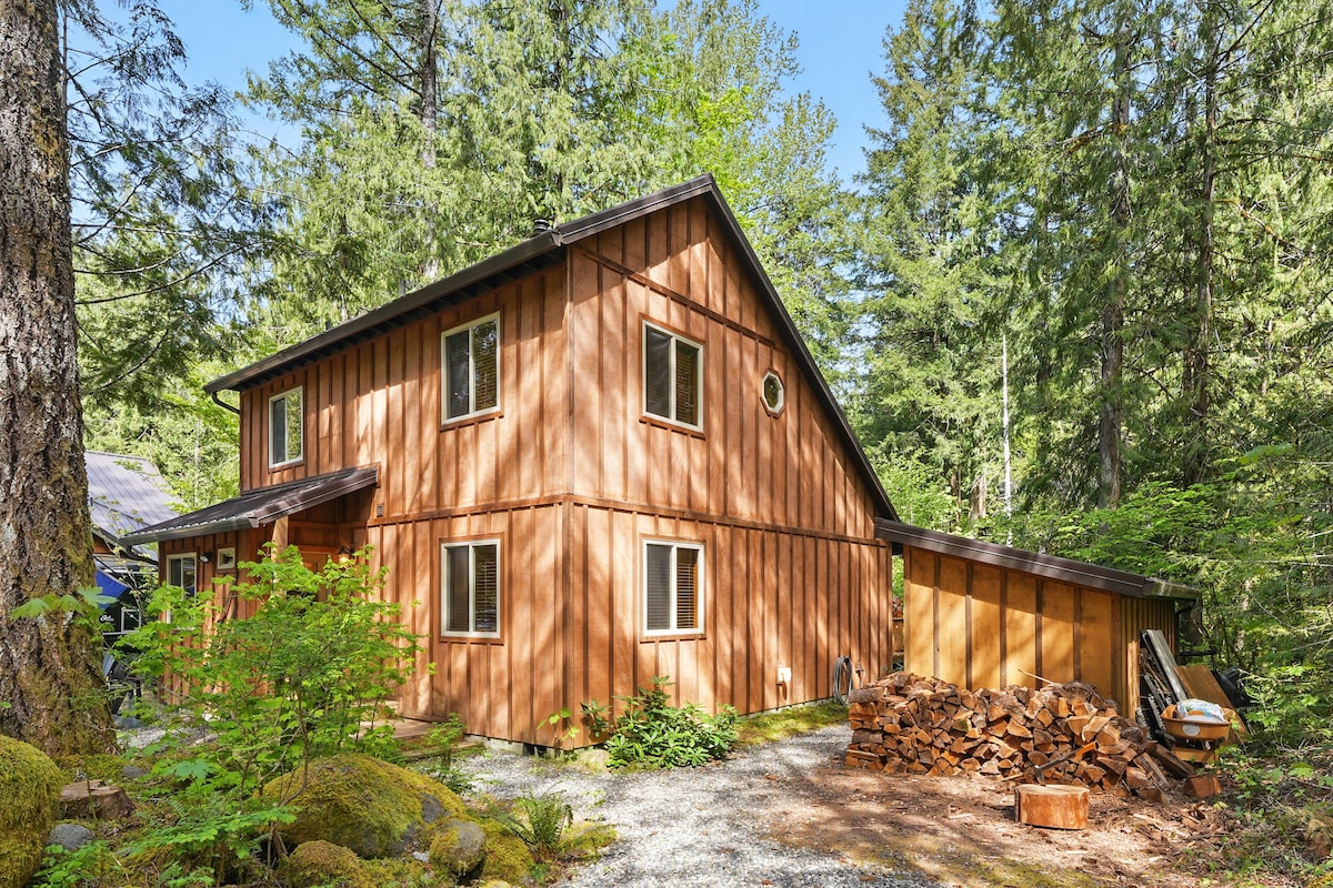 A wooden cabin sits amidst a lush forest, showcasing natural wood siding and a sloped roof. Windows allow views of the surrounding greenery. A stack of firewood is neatly arranged beside the cabin, with gravel pathways leading to the entrance.