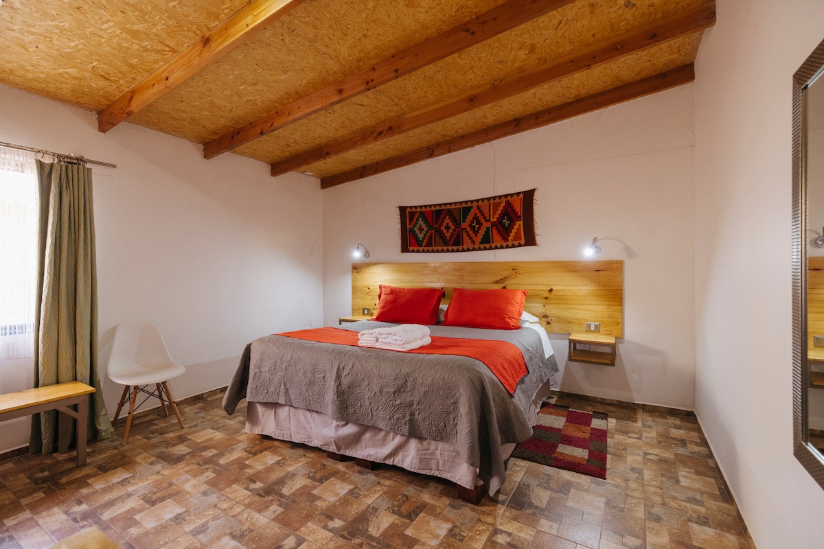 A spacious sleeping area features a large bed with colorful pillows and blankets. Wooden beams create warmth in the ceiling, and a textile piece adds an artistic touch above the headboard. Natural light enters through a window covered with light curtains, illuminating the cozy space.