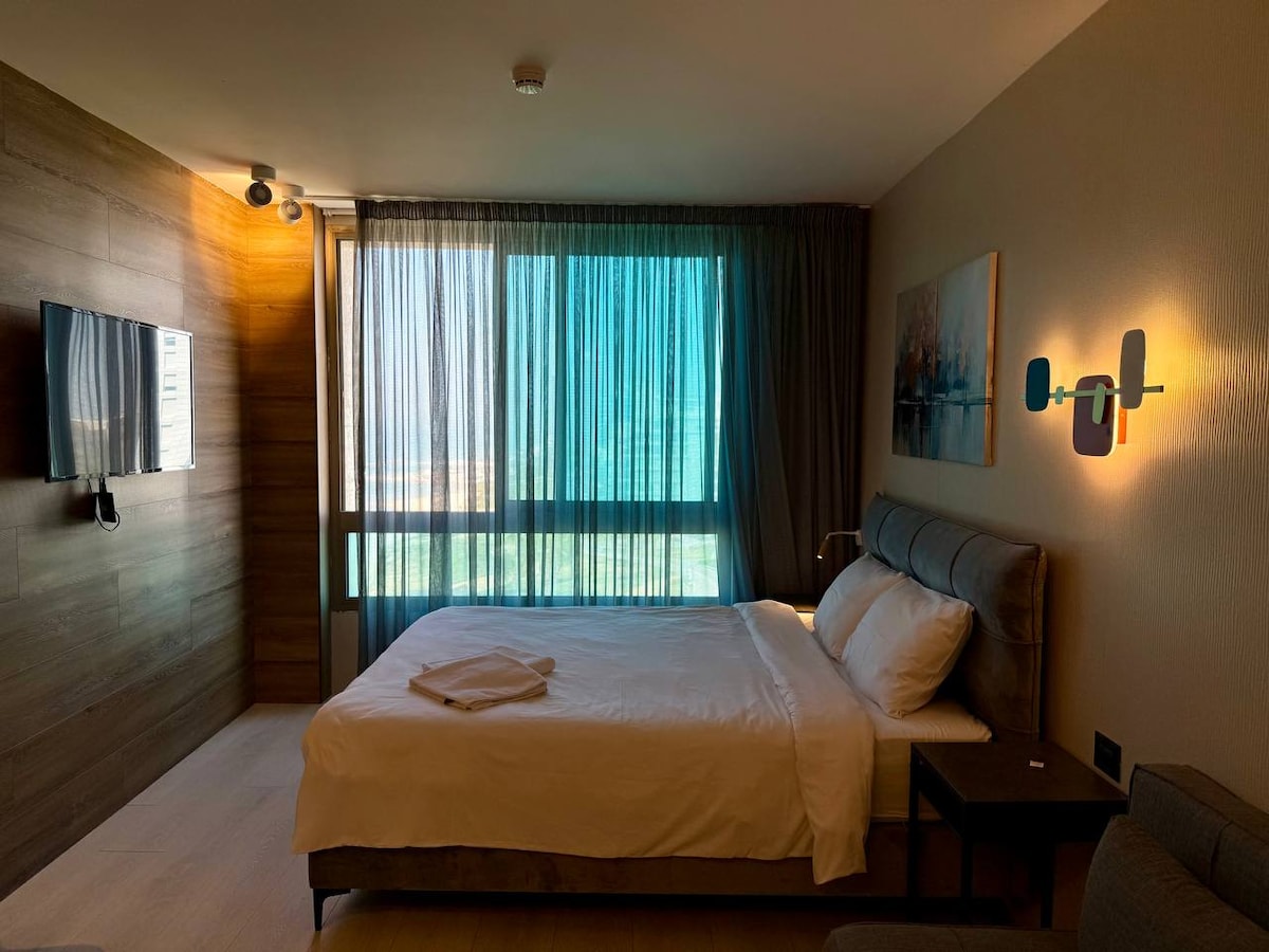 A well-appointed sleeping area features a spacious bed with crisp linens. Floor-to-ceiling windows allow for natural light, complemented by sheer curtains that softly diffuse brightness. A wall-mounted television is present, along with a contemporary piece of artwork that adds subtle color to the neutral decor.