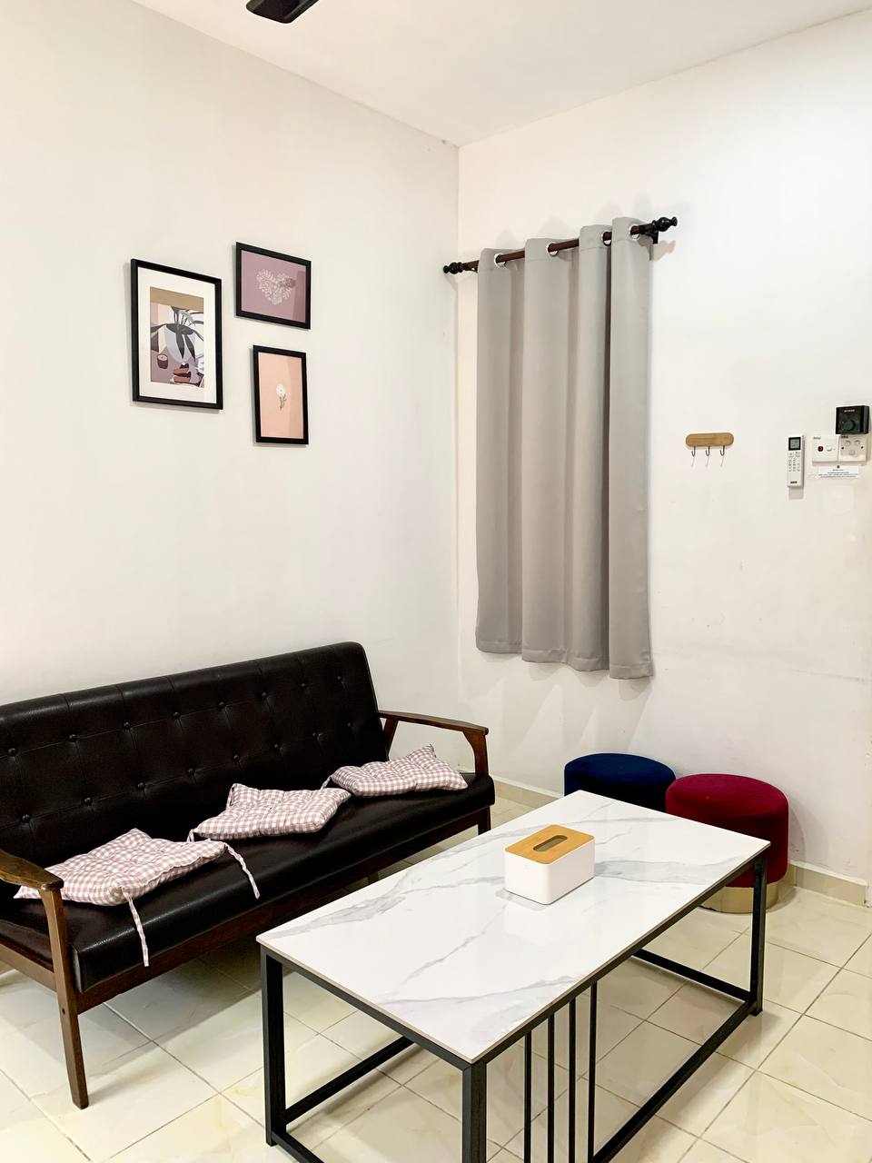 The seating area features a dark wooden sofa with patterned cushions and a marble-top coffee table. Framed wall art is displayed above the sofa. Two colorful poufs are positioned nearby, and light grey curtains frame the window, contributing to a relaxed environment.