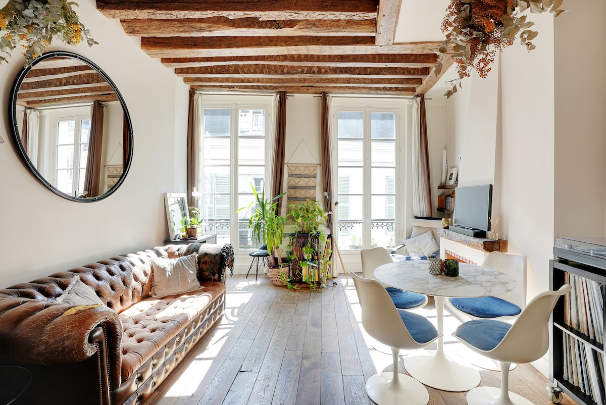 A living area is characterized by wooden beams overhead and a large circular mirror. A tufted sofa sits beside a round dining table with stylish chairs. Sunlight pours in through double windows, illuminating several potted plants and a television on a light-colored media console.