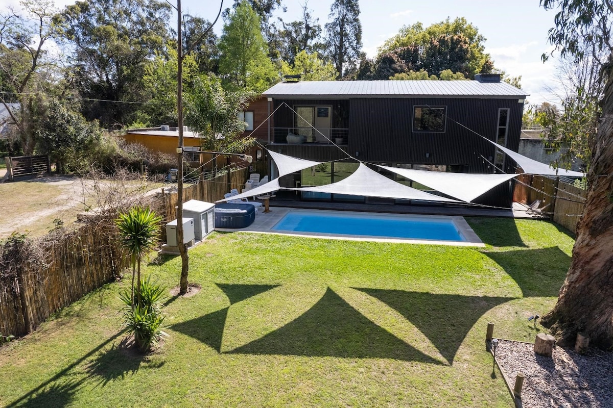 The outdoor area features a spacious green lawn bordered by trees. A swimming pool is centrally located, accompanied by a jacuzzi and shaded seating areas. The house is visible in the background, showcasing modern architectural elements amid a natural setting.